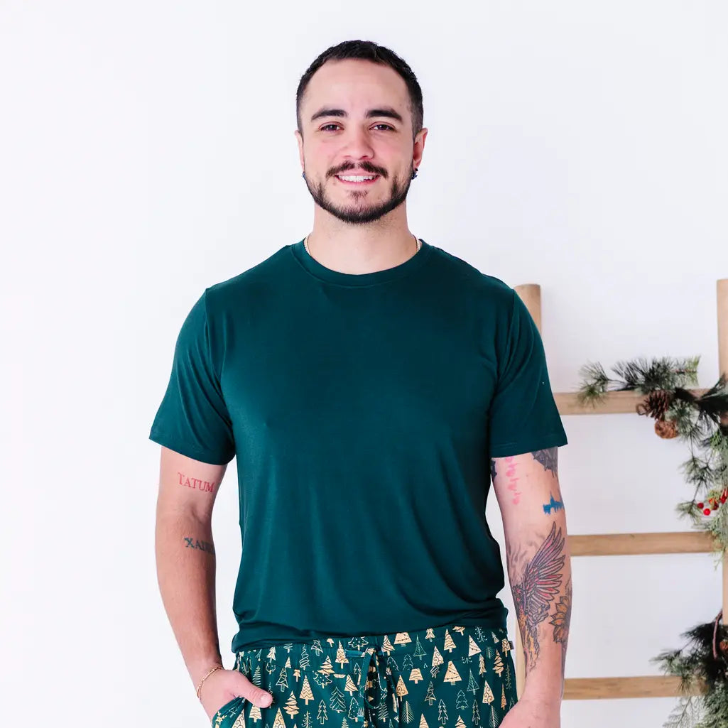A man smiling in a solid dark green crew neck tee, shown from the waist up with hands in the pockets of jogger pajama pants in a gold Christmas tree print.